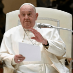 Pope Francis seated, holding notes and gesturing while speaking into a microphone during a public address at the Vatican.