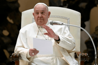 Pope Francis seated, holding notes and gesturing while speaking into a microphone during a public address at the Vatican.