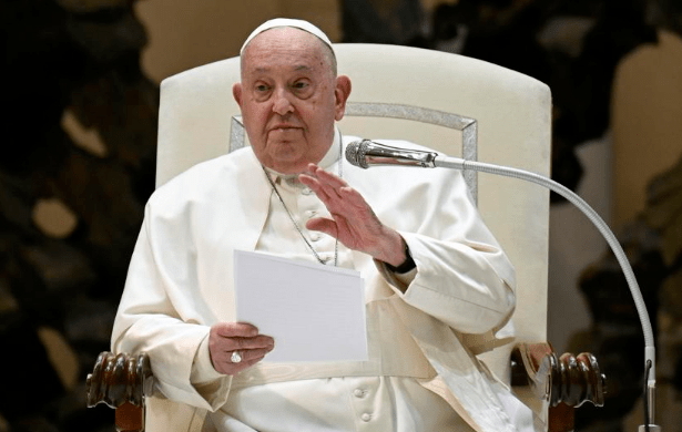 Pope Francis seated, holding notes and gesturing while speaking into a microphone during a public address at the Vatican.