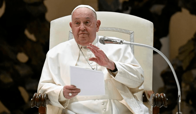 Pope Francis seated, holding notes and gesturing while speaking into a microphone during a public address at the Vatican.