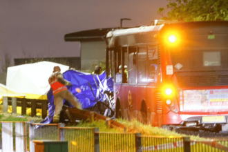 Emergency services at the scene of a fatal collision between a car and a bus on Bedfont Road in Feltham at night, with vehicles damaged and emergency lights flashing.
